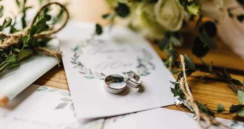 iStock-1455769561.jpg Stylish rings, flowers on wooden table background. Letters from the bride and groom. Vows. Engagement. Luxury marriage and weddi