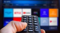 A man is holding a remote control of a smart TV in his hand. In the background you can see the television screen with streaming